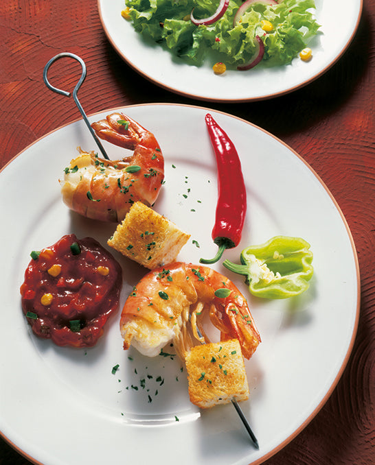 Scampi angerichtet als Spieß mit Brot. Auf dem Teller ist noch eine Chili und Paprika