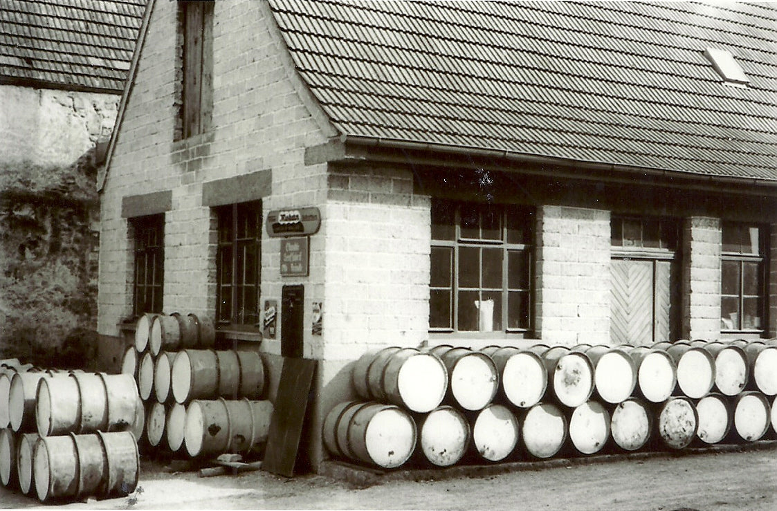 Oil barrels are stacked in front of an old brick house with transom windows. 