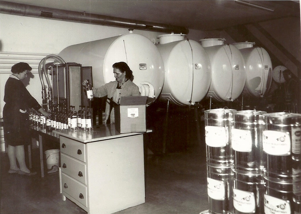 In front of white steel tanks, two women fill cooking oil into bottles by hand.   Jugs stand in front.